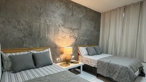 Habitación de hotel con dos camas, una pared de acento de concreto, una mesita de noche con lámpara y una ventana con cortinas.