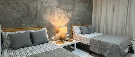 Habitación de hotel con dos camas, una pared de acento de concreto, una mesita de noche con lámpara y una ventana con cortinas.