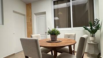 A dining area with a round wooden table, four chairs, a hanging light fixture, and a window with blinds.