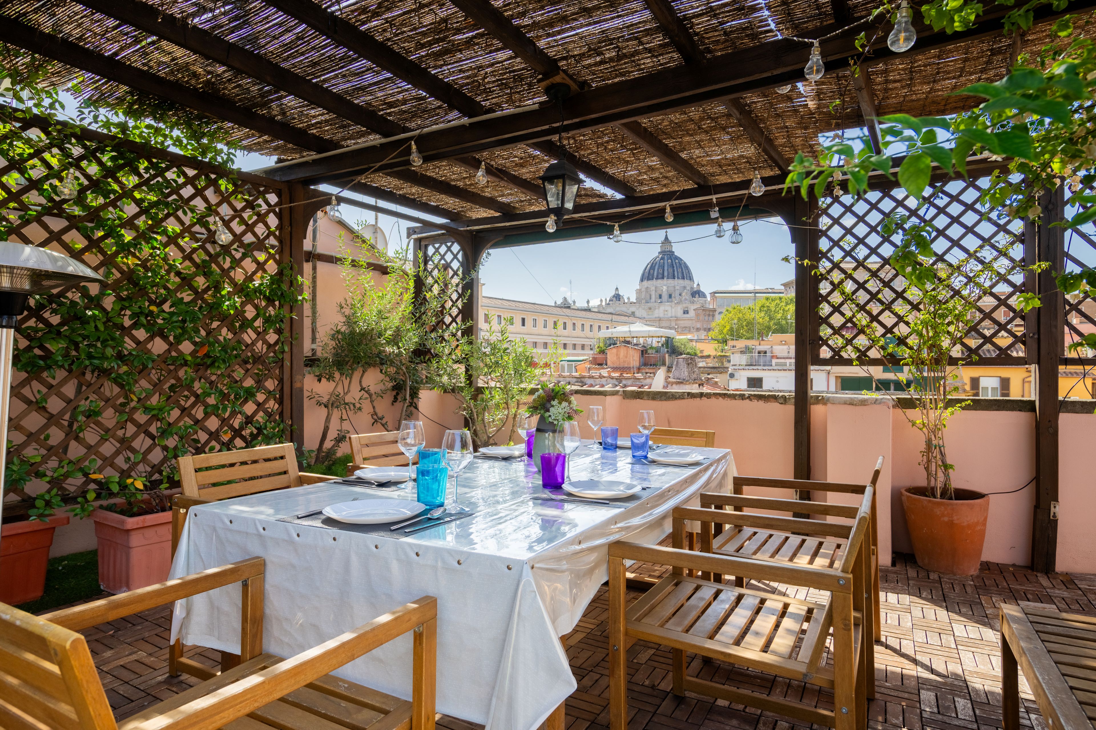 An outdoor dining area with a table set for two, surrounded by wooden chairs, under a pergola with a view of a cityscape and a dome structure.