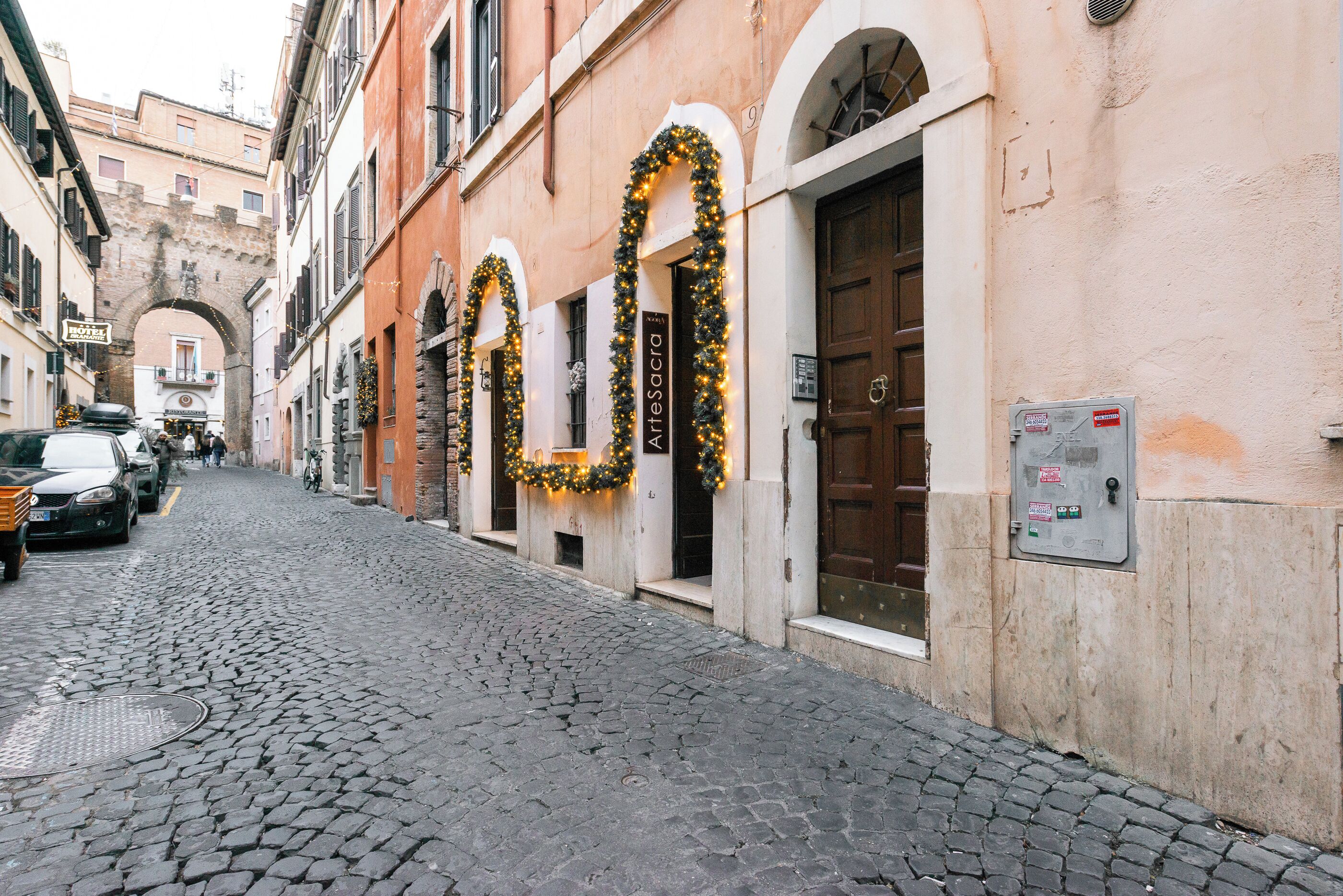 A cobblestone street with buildings, a sign for "Hotel Arte Sacra," and a brown door.