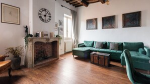 Een woonkamer met een open haard, een groene bank, houten vloeren en een klok aan de muur.