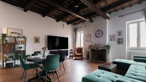 Een ruime woonkamer met een open haard, houten balken, een eethoek met groene stoelen en een televisie.