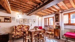 Una habitación tradicional con vigas de madera a la vista, un sofá floral, una mesa de comedor de madera con sillas, un banco rojo y una ventana con cortinas.