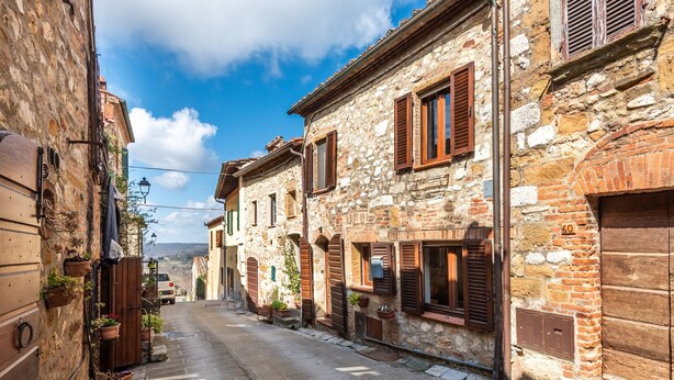 Una estrecha calle flanqueada por edificios de piedra, algunos con contraventanas de madera, y plantas en macetas.