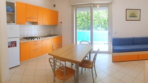 Een kleine keuken met oranje kastjes, een eettafel met stoelen en een blauwe bank in een lichte kamer met een groot raam.