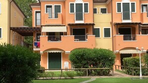 A multi-story residential building with balconies, a covered patio, and a well-maintained garden. - Coastal apartment with shared pool (Bibione)