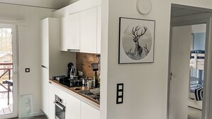 Eine moderne Küche mit weißen Schränken, einem Herd und einem Spülbecken. An der Wand hängt ein Bild von einem Reh und eine Tür führt in einen Raum mit Etagenbetten.
