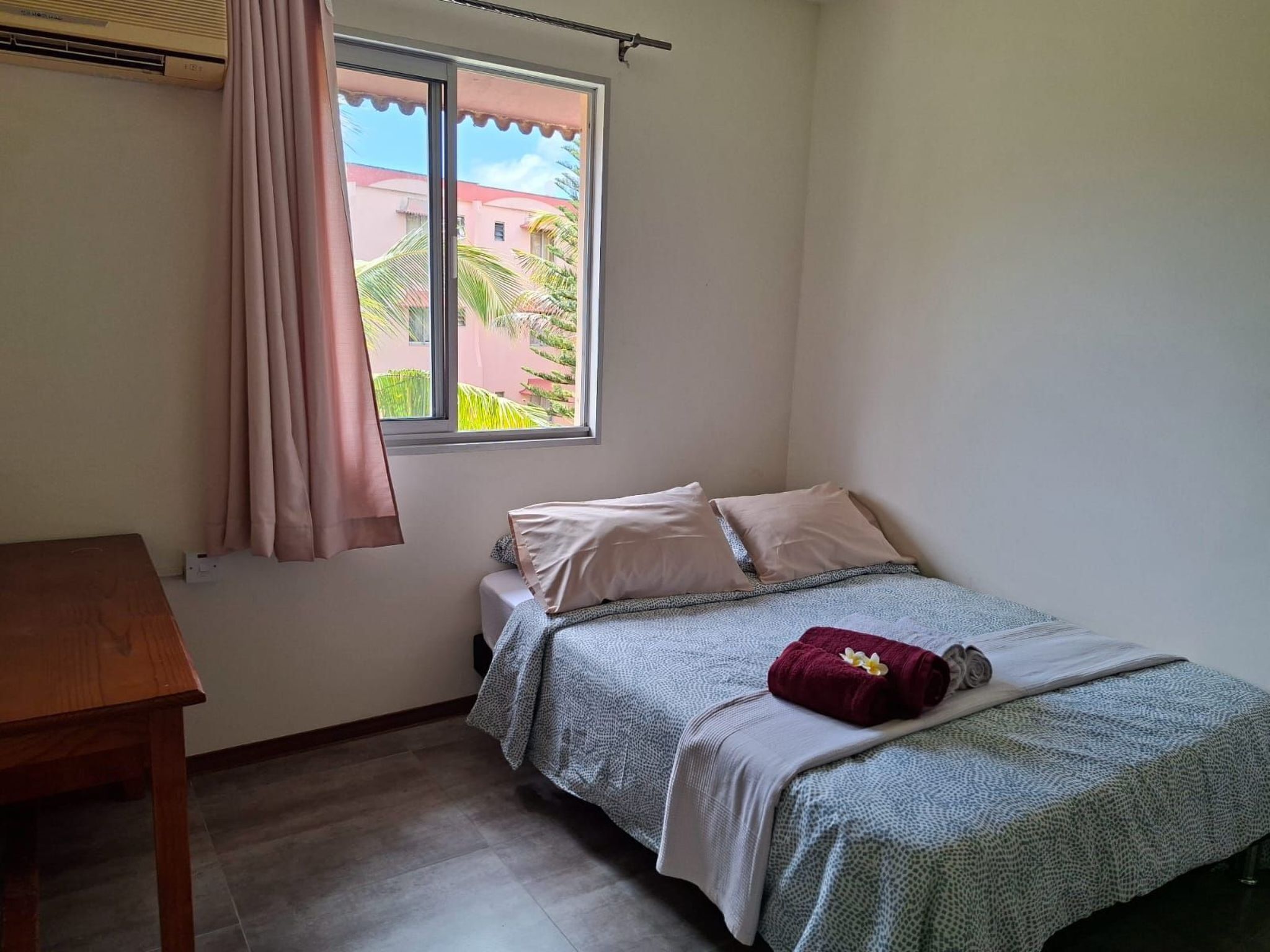 Een kleine, schone kamer met een enkel bed, een houten nachtkastje en een raam met uitzicht op gebouwen en groen.