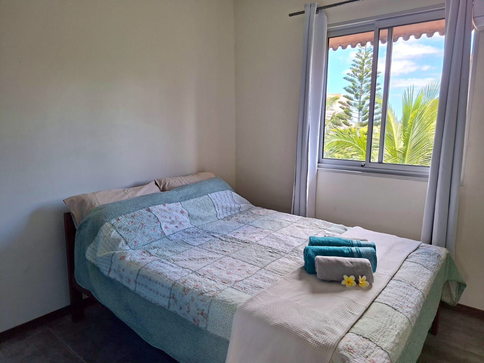 A neatly made bed with a floral-patterned bedspread and two towels, a window with a view of greenery, and a curtain.