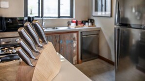 Een keuken met een houten snijplank met messen, een fornuis, een koelkast en een raam met jaloezieën.