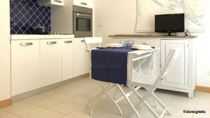 Ett kompakt kök med vita skåp, en spis och en mikrovågsugn. Där finns ett matbord med en blå duk och en stol. En tv är monterad på väggen ovanför bordet.