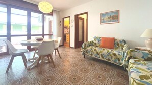 Un salon avec un sol à motifs, un canapé recouvert d’un tissu à fleurs, un coin repas avec une table et des chaises, et une cuisine visible à travers une porte ouverte.