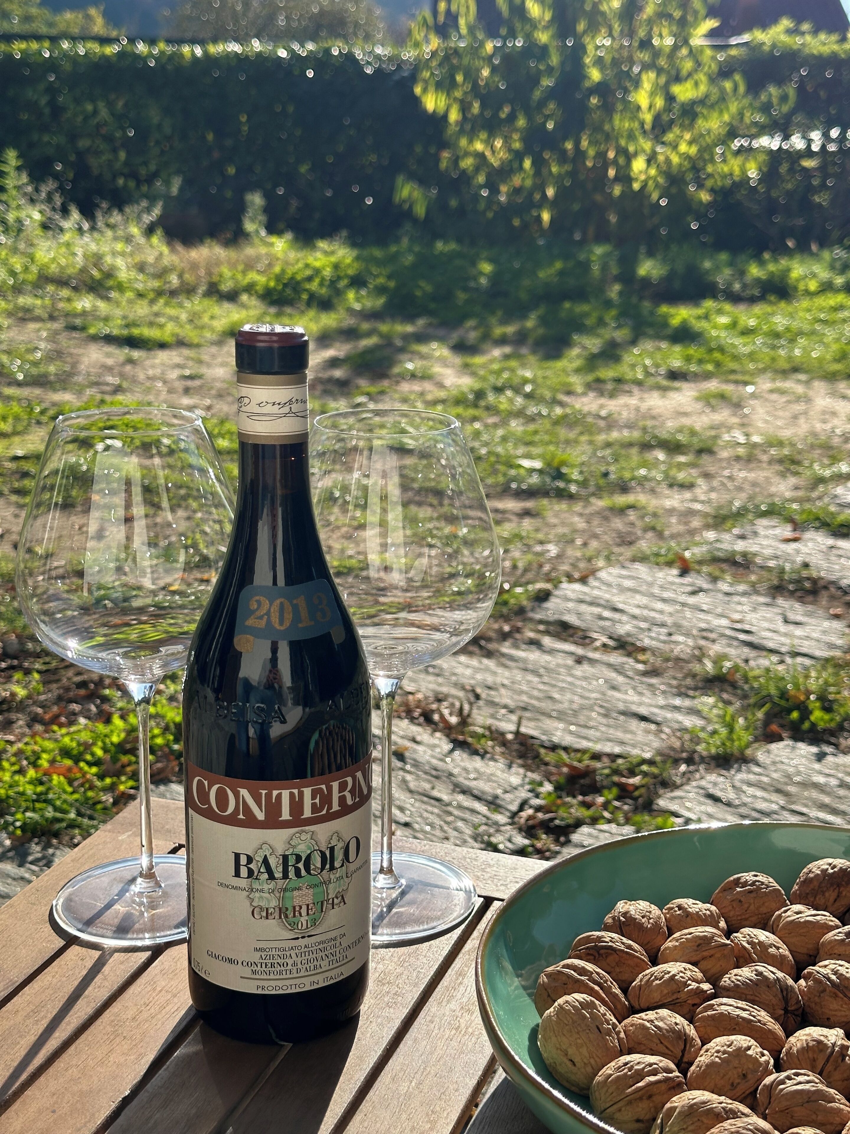 Eine Flasche Barolo Wein mit zwei leeren Weingläsern auf einem Holztisch, draußen im Freien. Im Hintergrund eine Schale mit Walnüssen und Grünpflanzen.