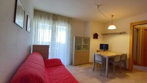A compact living space with a red sofa, a dining table with chairs, a television, and a small cabinet. - Family friendly vacation apartment with a balcony (Grado)