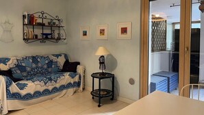 Een woonkamer met een gestreept bankje, een bijzettafeltje met een lamp, een boekenplank en een schuifdeur naar een balkon.