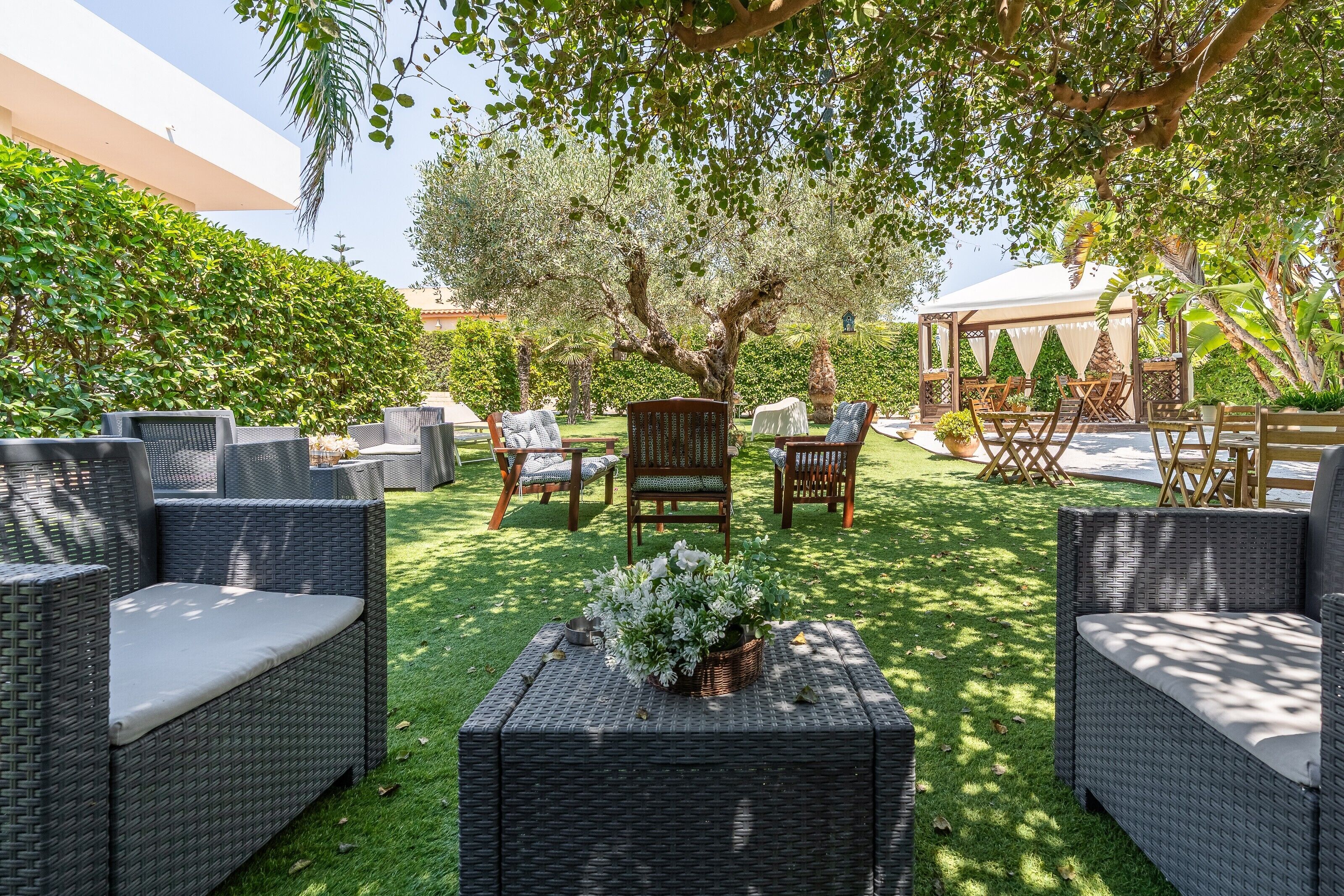 An outdoor seating area with wicker furniture, a central tree, and a covered structure in the background.