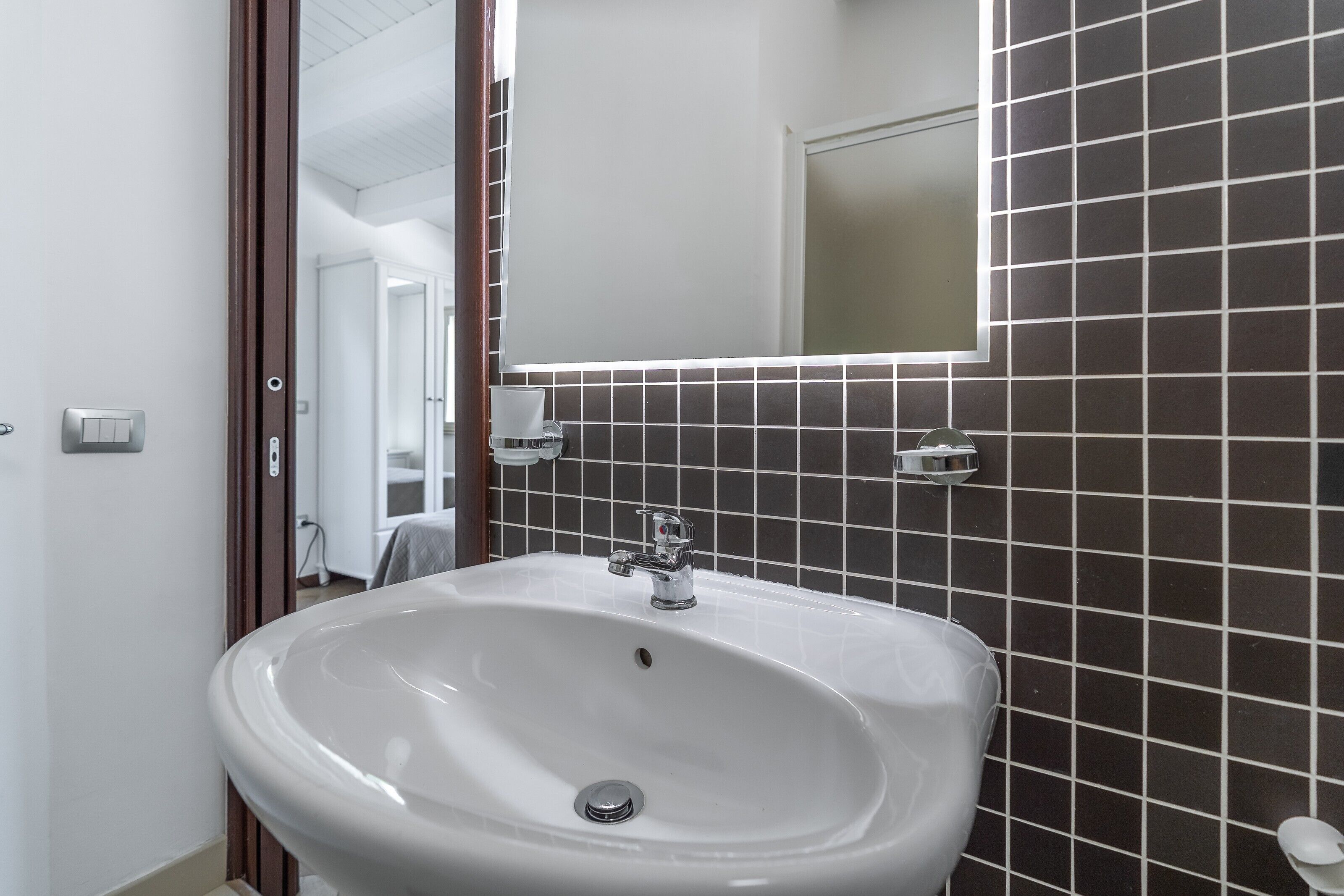 Une salle de bain avec un lavabo blanc, des murs recouverts de carreaux foncés et un miroir.