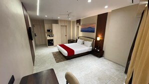 A hotel room with a bed, a desk, a chair, a nightstand, a lamp, and a painting on the wall. - Hotel Saffron Suites (Chaibasa)