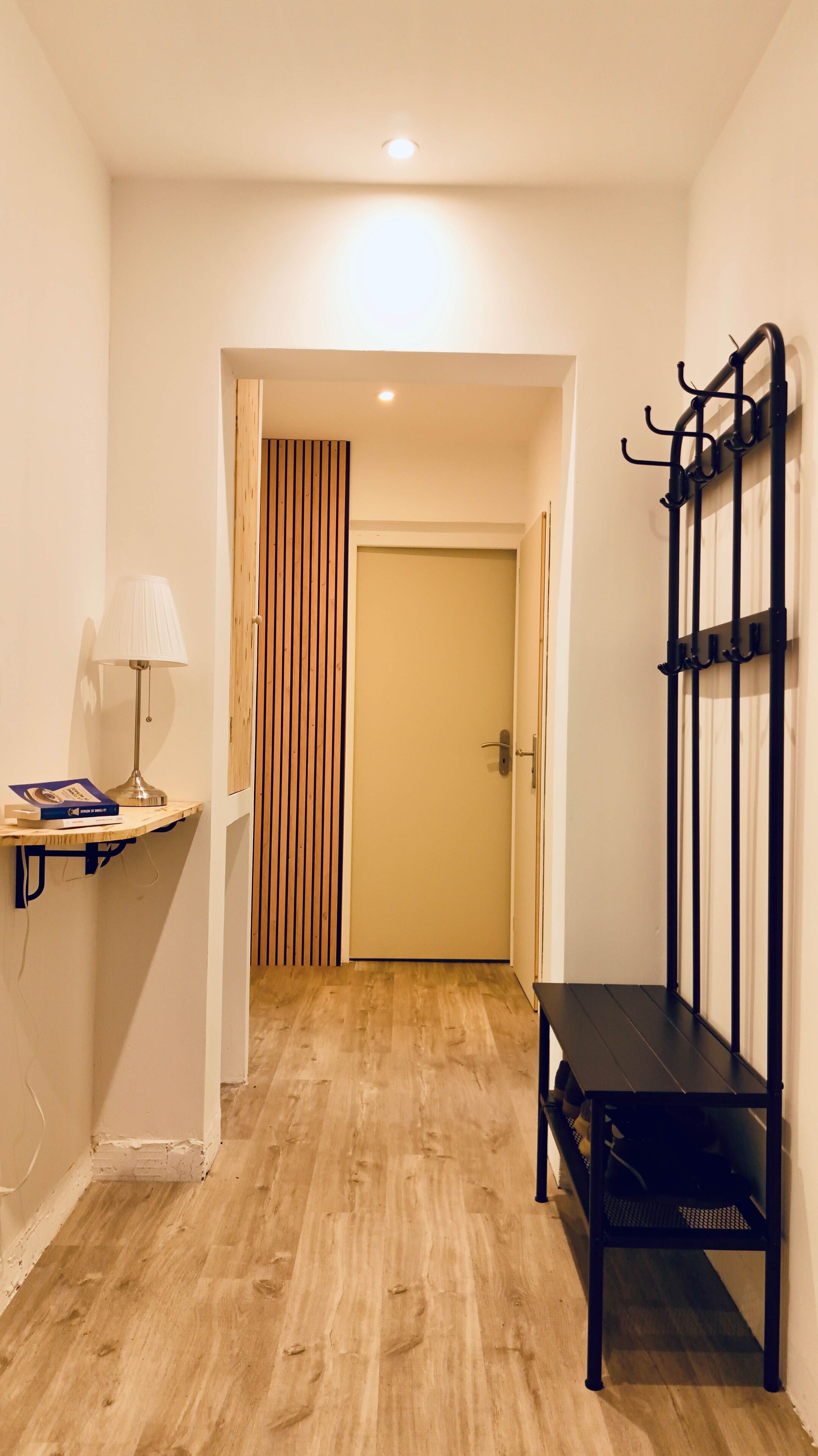 A hallway with a wooden floor, a black bench, a wall-mounted coat rack, and a door with vertical blinds.