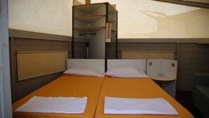 Habitación con dos camas, cada una con una almohada blanca y una toalla blanca doblada. Encima de las camas hay un mueble estante de madera.