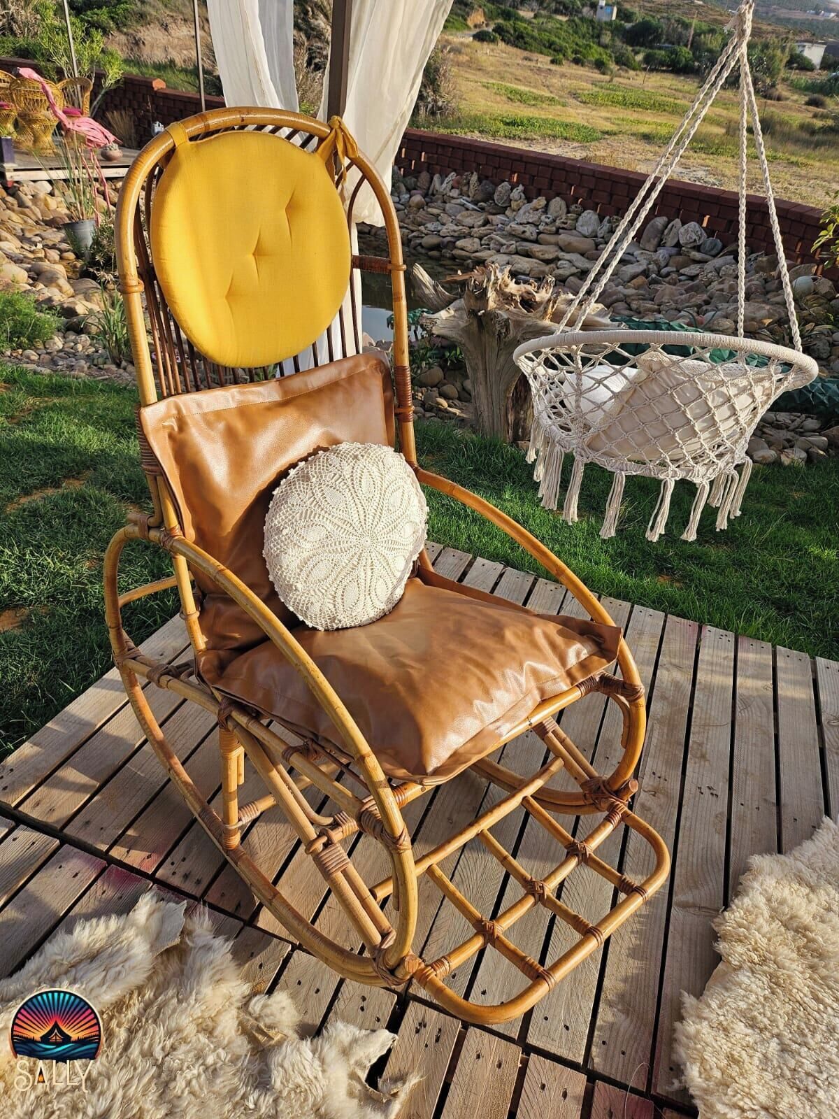 Una mecedora de mimbre con un cojín amarillo y una almohada blanca, sobre una terraza de madera con un paisaje de césped al fondo.