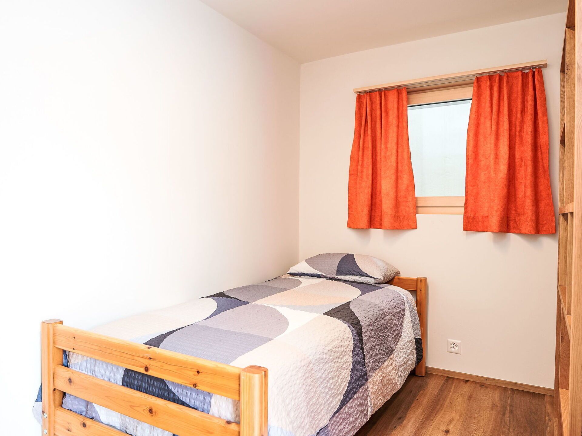 Une petite chambre avec un lit simple, une tête de lit en bois, une fenêtre avec des rideaux orange et un mur blanc uni.