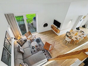 Ein modernes Wohnzimmer mit Sofa, Sesseln, einem Esstisch für sechs Personen, einem Fernseher und einer Glasschiebetür zu einem Außenbereich.