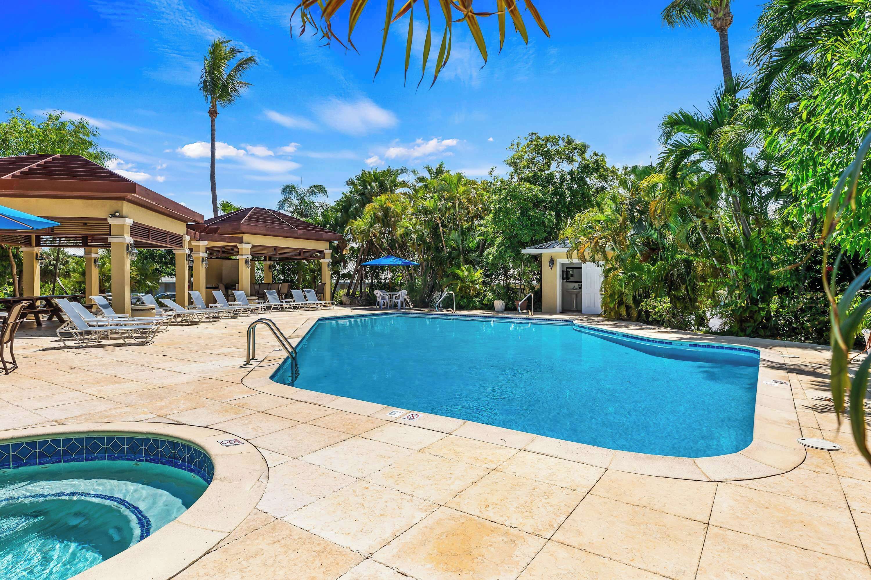 Piscina com espreguiçadeiras e hidromassagem, cercada por palmeiras e estruturas cobertas.