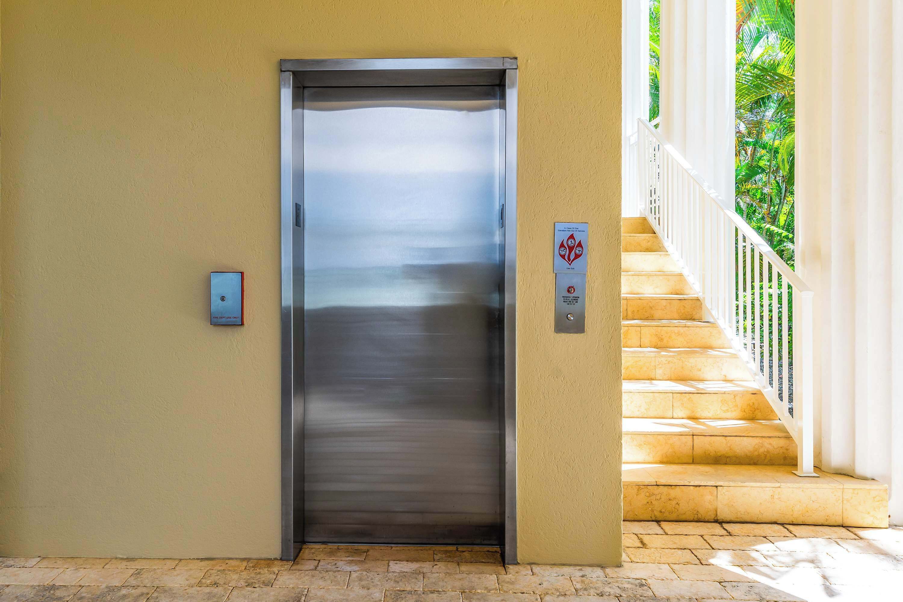 Un ascenseur aux finitions métalliques, un escalier avec des rampes blanches et un panneau indiquant les consignes de sécurité en cas d’urgence.