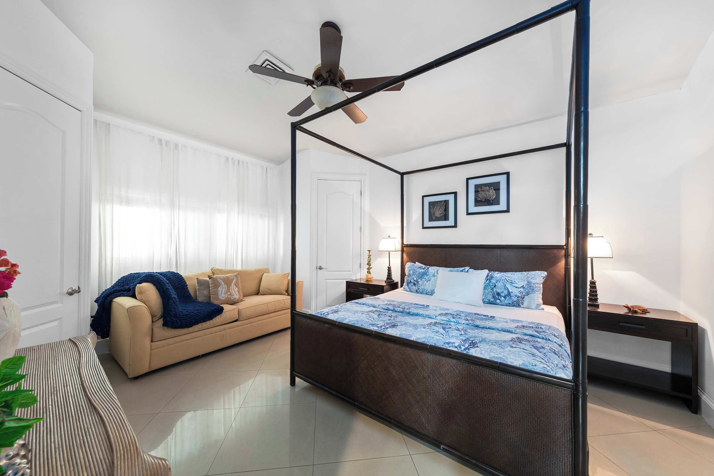 Ein Bett mit Baldachin, ein Deckenventilator, ein beiges Sofa und ein Nachttisch mit Lampe.