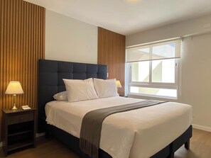 A hotel room with a large bed, wooden paneling on the walls, a window with blinds, and a nightstand with a lamp. - w Marvelous 2BR w Balcony in Miraflores (Lima)
