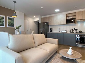 A modern living room with a beige sofa, a round wooden coffee table, and a kitchen area with grey cabinets and stainless steel appliances. - w Marvelous 2BR w Balcony in Miraflores (Lima)