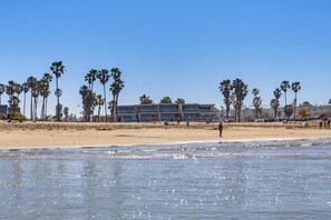 A beach with palm trees, a building with large windows, and people on the sand. - 6BR/6BA Luxury Group-Friendly (San Diego)