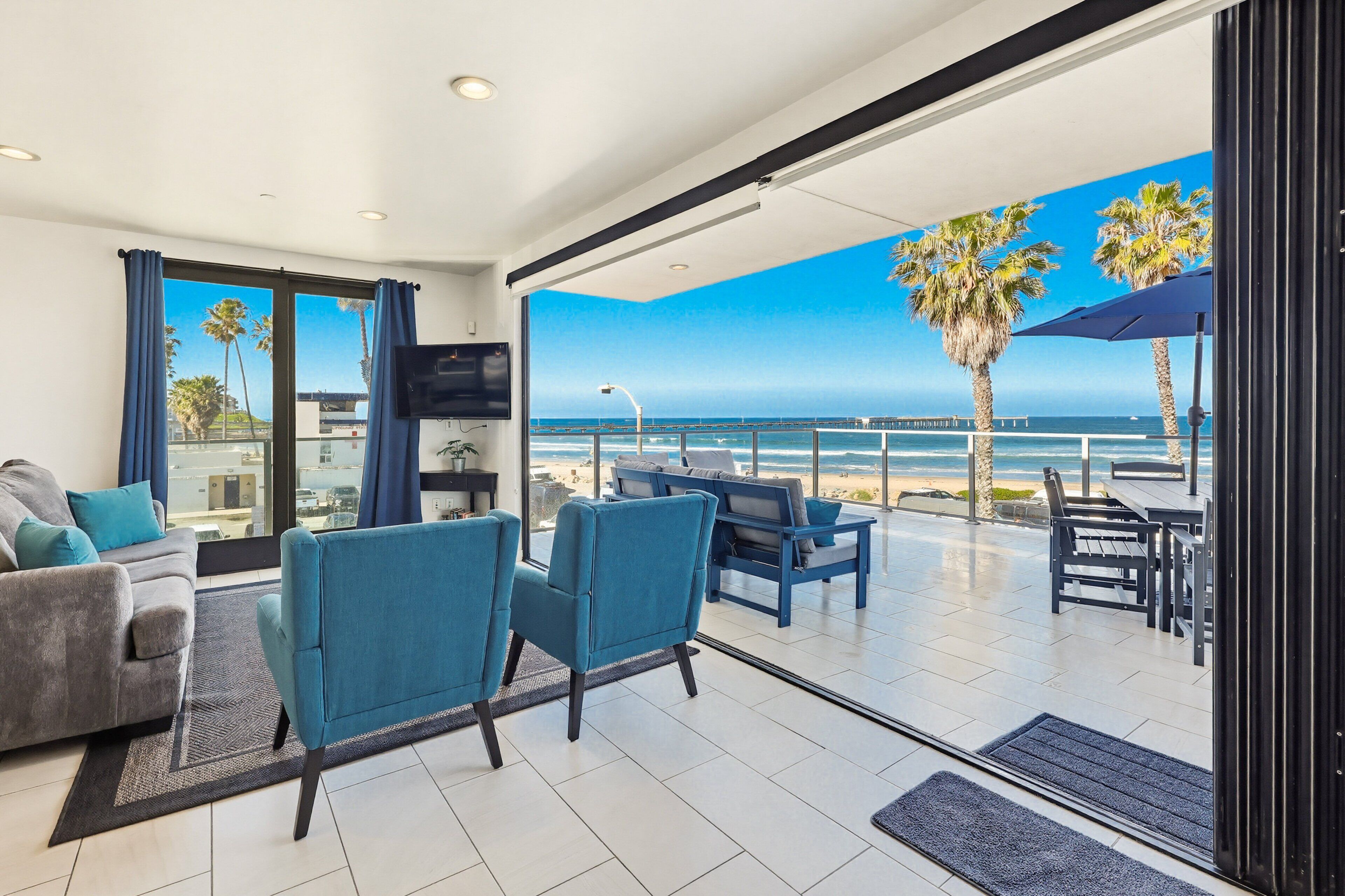 Ein modernes Wohnzimmer mit einer großen Schiebeglasdoor zum Balkon mit Blick auf das Meer und Palmen.