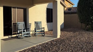 Un patio cubierto con dos sillas, una mesa, una puerta que da a un espacio interior y un pequeño jardín con grava.