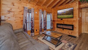 Ein Holzblockhaus-Interieur mit einer Couch, einem an der Wand montierten Fernseher und Vorhängen an den Fenstern.