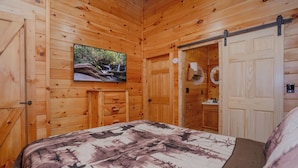 Ein Schlafzimmer einer Holzhütte mit einem Bett, einem an der Wand befestigten Fernseher und einer Tür zu einem Badezimmer.