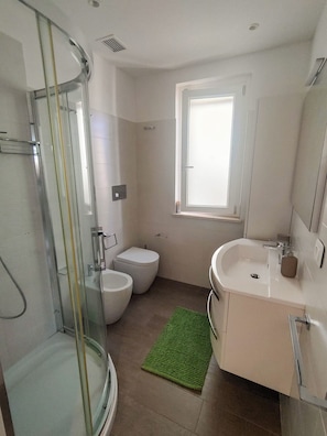A modern bathroom with a glass shower enclosure, a toilet, a sink, and a window providing natural light. - Villino degli Ulivi (Bergeggi)