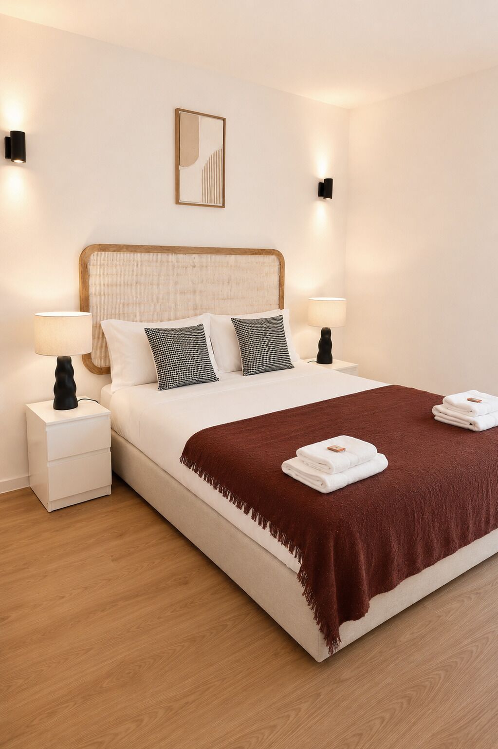 Een net opgemaakt bed met een bruine plaid en witte kussens, twee nachtlampjes en een wandlamp.