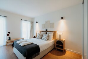 A modern bedroom with a large bed, bedside lamps, a wooden bench, and a bedside table with a vase of flowers. - Anjos Apartments (Lisbon)