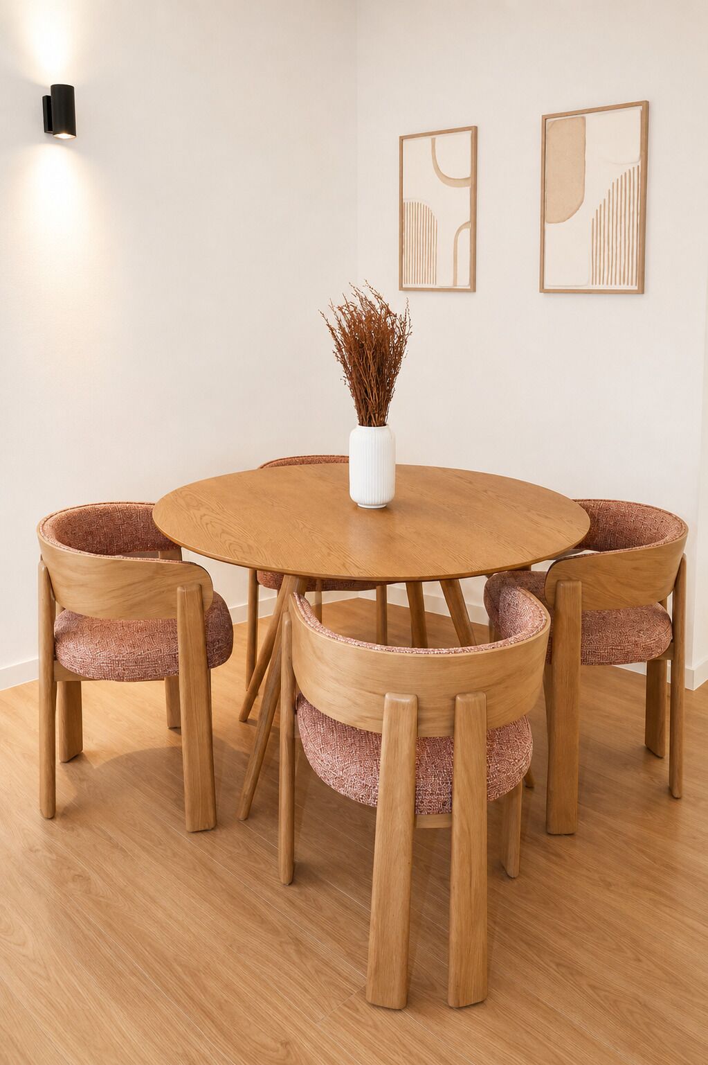 Een ronde houten eettafel met zes stoelen, een witte vaas met gedroogde planten, en twee abstracte kunstwerken aan de muur.