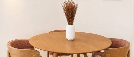 Een ronde houten eettafel met zes stoelen, een witte vaas met gedroogde planten, en twee abstracte kunstwerken aan de muur.