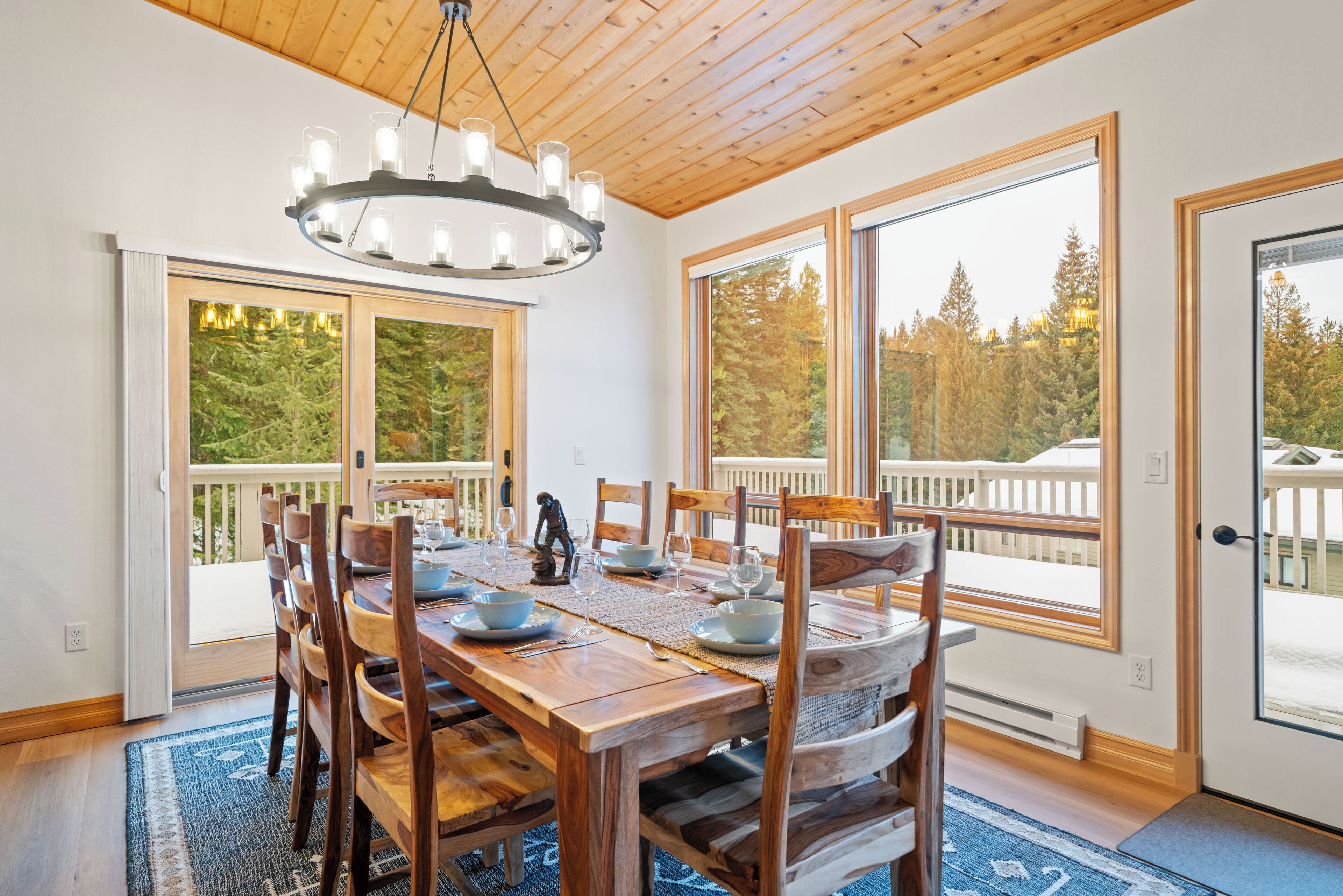 Un comedor con una mesa de madera, sillas y una araña. Se ven grandes ventanales con vistas a árboles y una terraza al exterior.