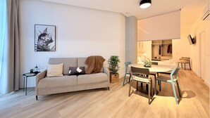 Uma sala de estar moderna com um sofá, uma mesa de jantar e cadeiras. Há uma mesinha lateral com uma garrafa e copos. Uma planta em vaso está no chão e um quadro na parede.
