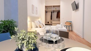 Quarto de hotel moderno com cama, mesa de cabeceira com copos de vinho, televisão na parede e armário com roupas penduradas.