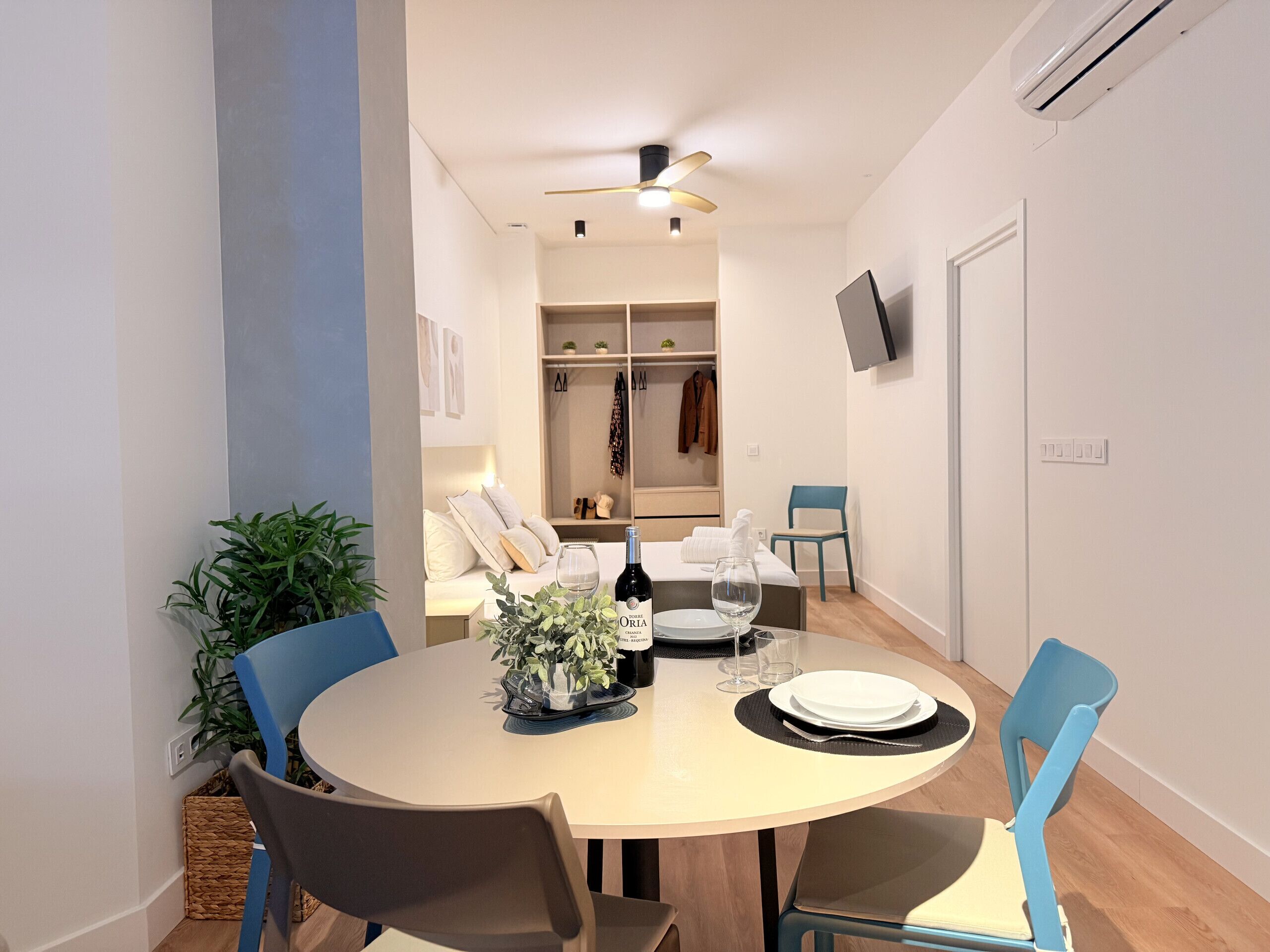 A dining area with a round table, chairs, and a ceiling fan. There is a TV mounted on the wall and a plant on the table.