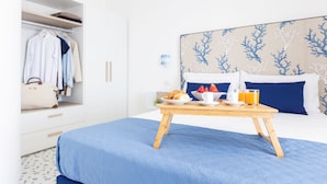 A neatly made bed with a blue bedspread, a wooden bedside table with breakfast items, a white wardrobe with hanging clothes, and a headboard with a blue and white coral pattern. - SEA FRONT TERRACE APARTMENT MARINA GRANDE SORRENTO (Sorrento)