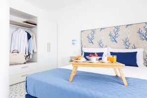 A neatly made bed with a blue bedspread, a wooden bedside table with breakfast items, a white wardrobe with hanging clothes, and a headboard with a blue and white coral pattern. - SEA FRONT TERRACE APARTMENT MARINA GRANDE SORRENTO (Sorrento)