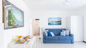A bright living room with a blue sofa, a dining table with breakfast, and a large painting of a coastal scene. - SEA FRONT TERRACE APARTMENT MARINA GRANDE SORRENTO (Sorrento)